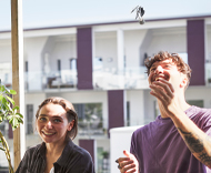 Lachendes Paar wirft Wohnungsschlüssel hoch, moderne Gebäude im Hintergrund.